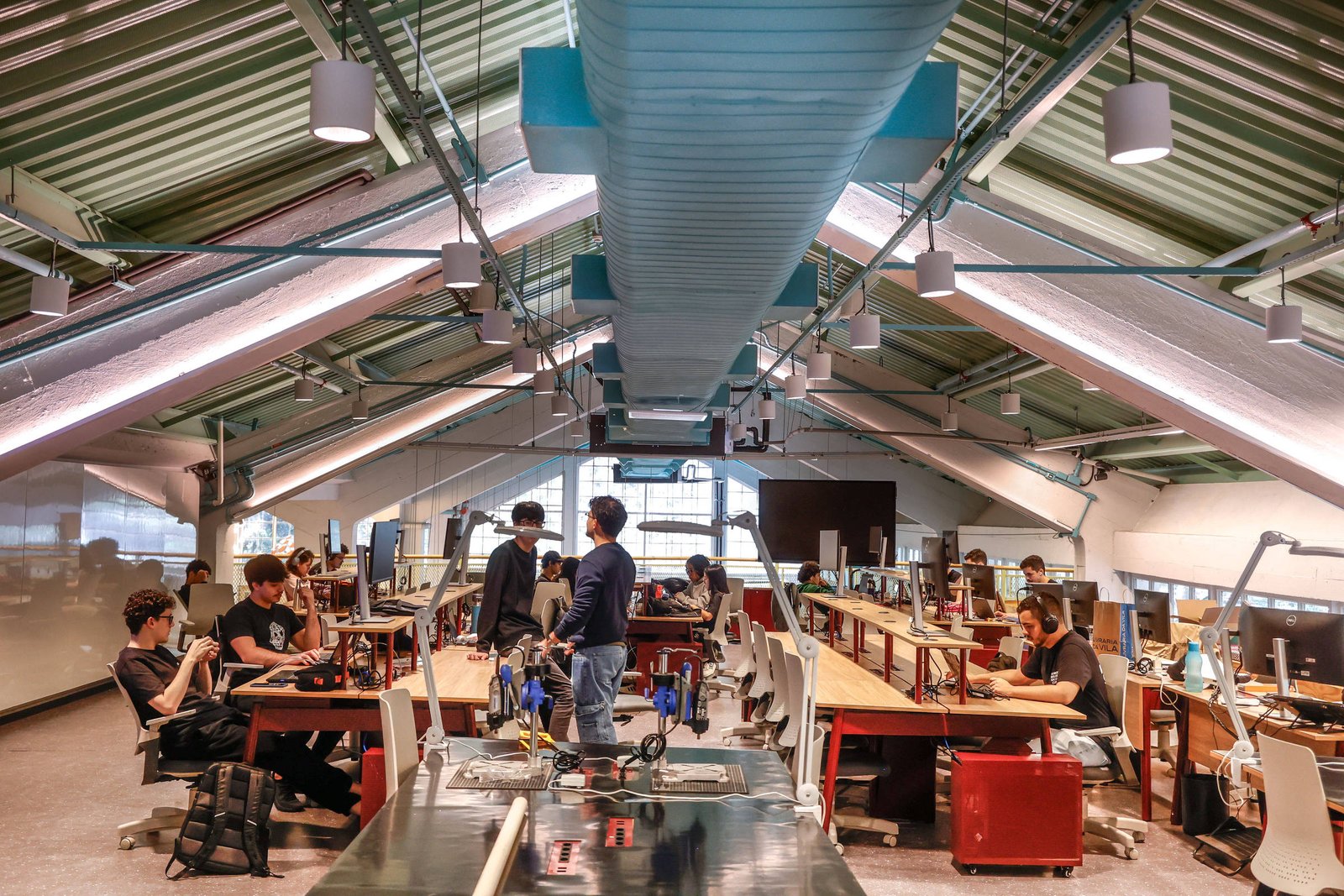Ambiente interno de laboratório de engenharia com teto inclinado e iluminação natural. Várias pessoas sentadas e em pé trabalham em bancadas com equipamentos eletrônicos e computadores. No centro, duas pessoas conversam próximas a uma mesa com componentes técnicos. O espaço é amplo, com cadeiras brancas e estrutura metálica aparente no teto.