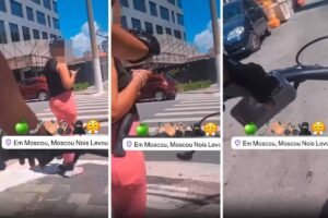 Mulher com blusa preta e saia rosa atravessa faixa de pedestres em rua de Moscou. Edifício moderno e carros estacionados ao fundo sob céu azul com poucas nuvens.