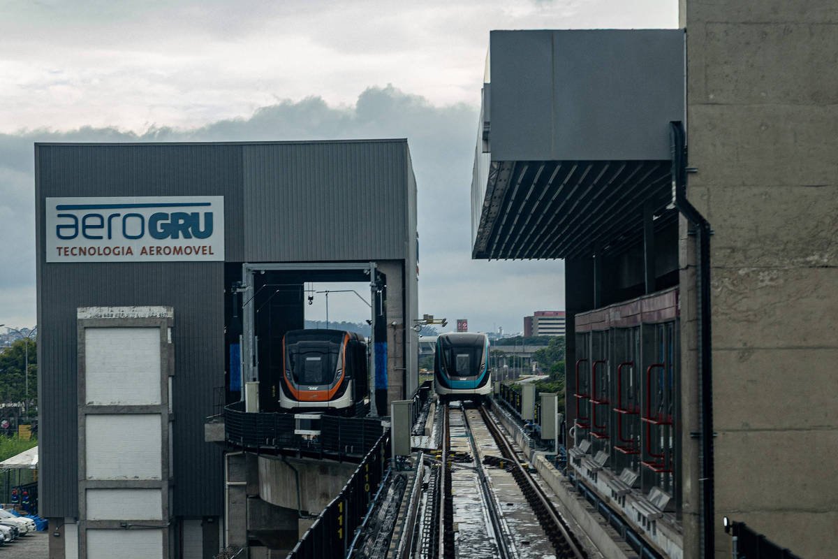 Dois trens do AeroGRU estão parados em uma estação elevada. A estrutura da estação é moderna, com trilhos elevados e sinalização ao fundo. O céu está nublado.
