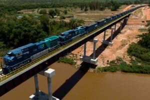 Trem de carga azul com várias locomotivas e vagões segue sobre ponte elevada de concreto que cruza rio de água barrenta. Vegetação densa e área rural cercam a ponte sob céu claro.