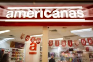 Fachada da loja Americanas com letreiro iluminado em vermelho e branco. No interior, decoração com letras vermelhas penduradas formando palavras, prateleiras com produtos e clientes circulando.