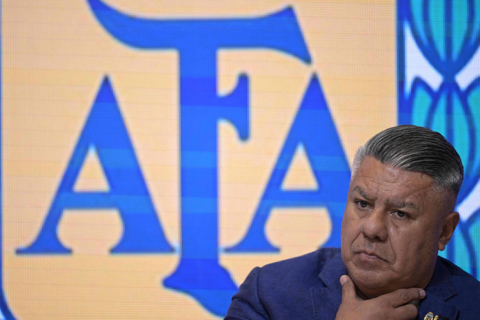 Homem de meia-idade com cabelo grisalho penteado para trás, vestindo terno azul, está com a mão no pescoço e expressão séria. Ao fundo, logo grande da AFA em azul sobre fundo amarelo.