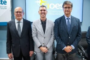 Três homens posam lado a lado em ambiente corporativo. O homem à esquerda veste terno preto com gravata cinza, o do centro usa terno cinza claro com camisa preta, e o da direita está com terno azul escuro e gravata azul. Ao fundo, tela exibe logotipo com texto relacionado à região metropolitana de São Paulo.