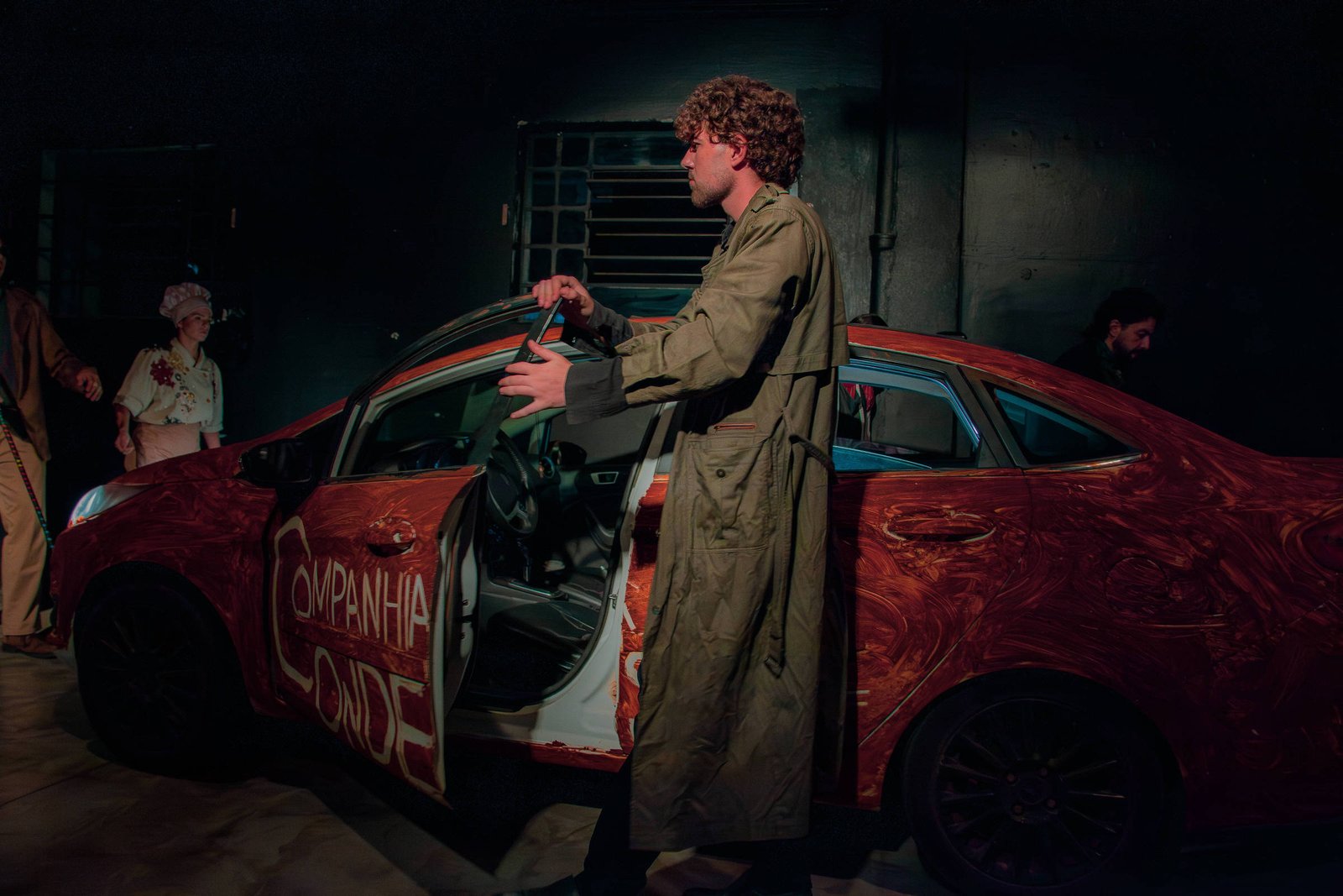 Homem de cabelos cacheados e sobretudo verde está ao lado de carro vermelho com porta dianteira aberta e inscrições brancas nas portas. Ambiente escuro sugere palco, com outras pessoas ao fundo parcialmente visíveis.