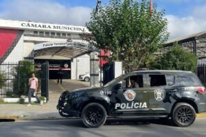 Viatura preta da polícia estacionada em frente à entrada da Câmara Municipal, com placa e logotipos visíveis. Pessoa caminha na calçada próxima ao portão de entrada. Árvore e céu parcialmente nublado ao fundo.