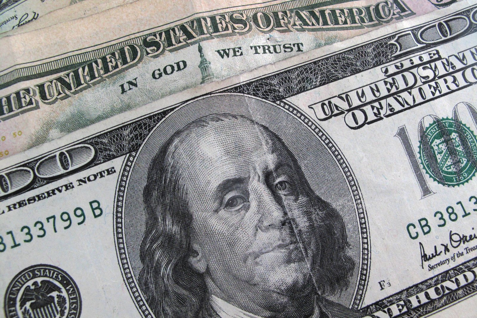 Notas de dólar americano sobrepostas, com foco na nota de cem dólares mostrando o retrato de Benjamin Franklin e detalhes do texto