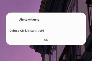 Tela de celular mostra alerta externo da Defesa Civil sobre fundo de céu roxo ao entardecer. Parte superior de prédio antigo com janelas e sacada aparece à direita.