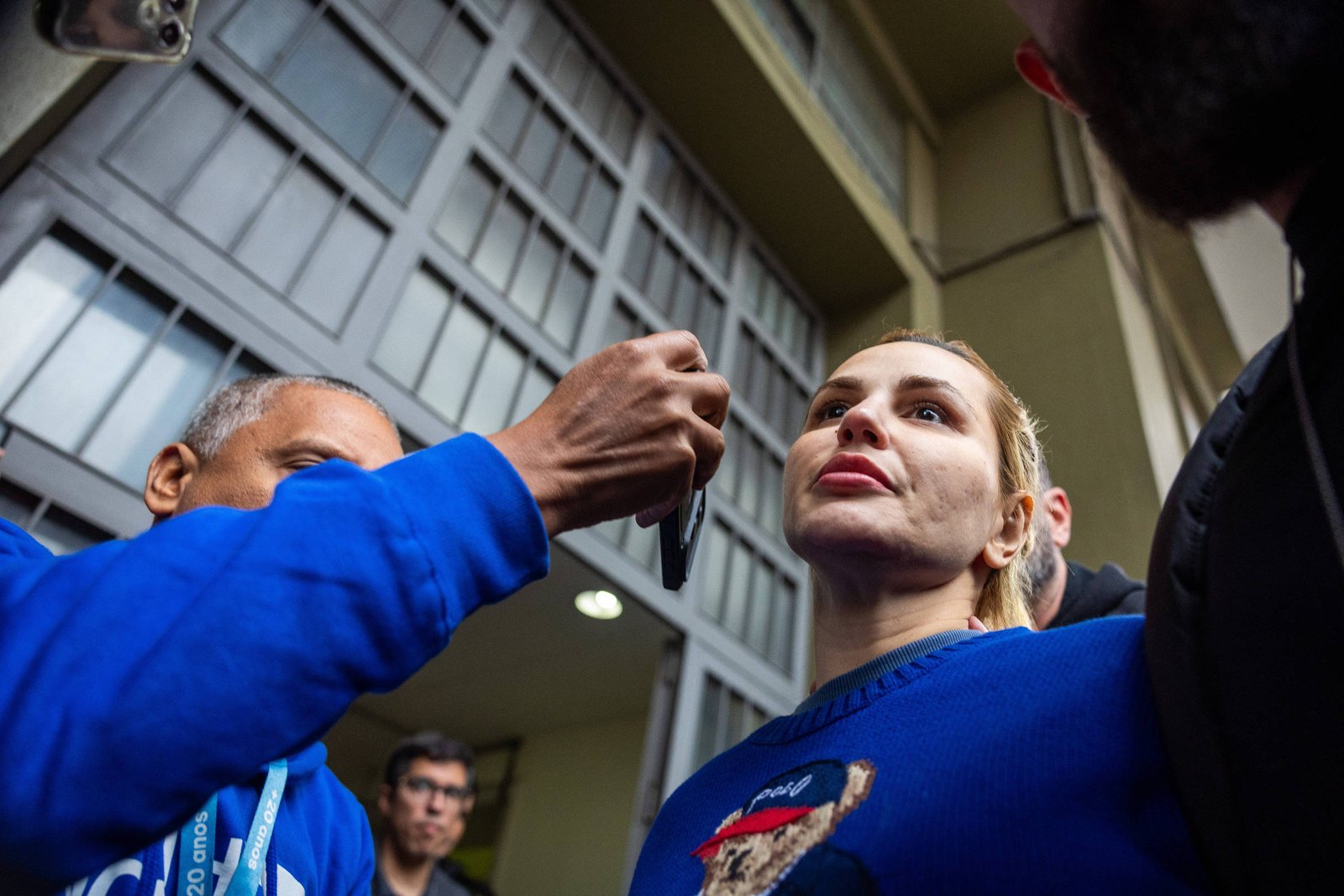 Mulher loira com suéter azul é entrevistada por homem de pele escura e jaqueta azul, que segura um microfone próximo ao rosto dela. Ao fundo, outras pessoas observam em frente a uma estrutura de janelas metálicas.