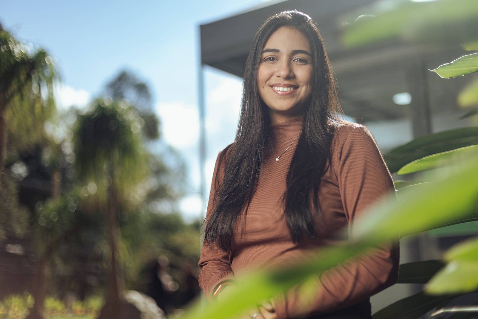 Mulher jovem com cabelo longo e escuro usa blusa marrom e sorri para a câmera. Ela está em área externa com plantas e árvores ao fundo, sob luz natural do dia.