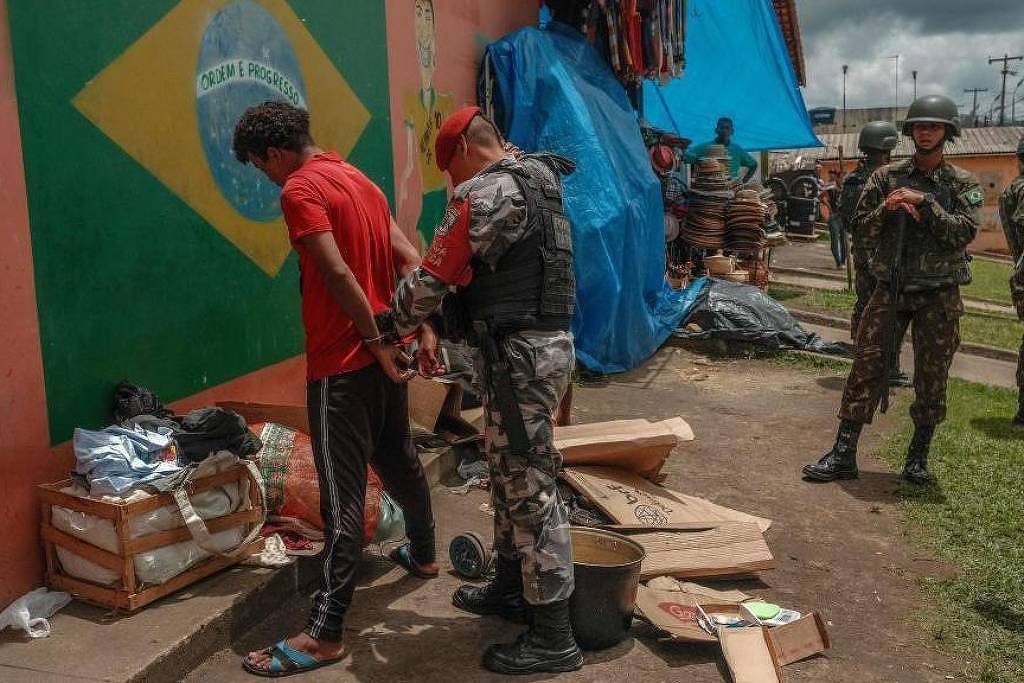 Policial prende homem de camiseta vermelha e calça preta com as mãos para trás em frente a muro pintado com a bandeira do Brasil. Outros militares estão próximos, alguns observando a cena, em área externa com barracas e objetos espalhados.