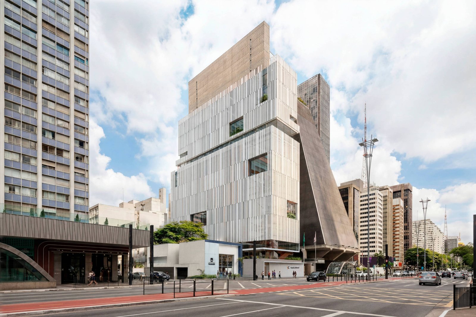 Prédio alto com fachada em painéis verticais claros e estrutura angular escura em destaque, localizado em avenida movimentada com trânsito e pedestres. Edifícios residenciais e comerciais cercam a construção sob céu parcialmente nublado.