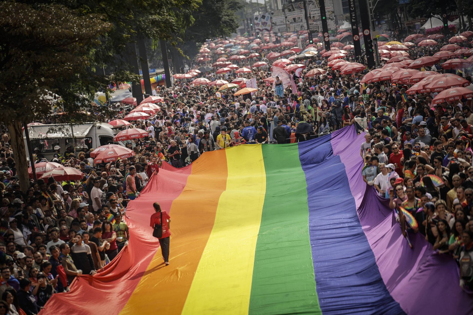 A imagem mostra uma grande multidão reunida em uma rua durante uma celebração do Orgulho LGBTQ+. No centro, uma enorme bandeira do arco-íris é estendida, enquanto pessoas caminham ao lado dela. Ao fundo, há muitas pessoas com guarda-sóis vermelhos, e a atmosfera é festiva e colorida.