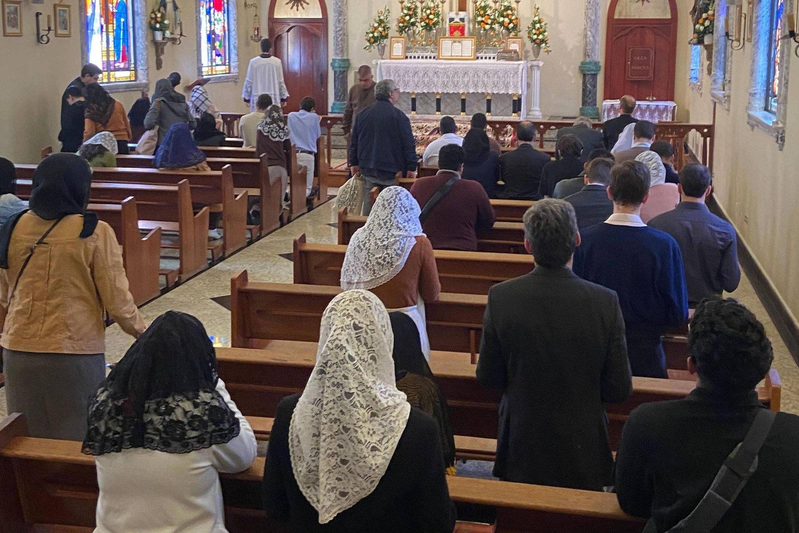 Pessoas sentadas e em pé nos bancos de madeira de uma igreja, algumas mulheres com véus brancos ou pretos na cabeça, voltadas para o altar decorado com flores e velas. Janelas com vitrais coloridos iluminam o ambiente interno.
