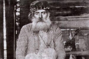 Homem idoso com barba e cabelo longos, usando faixa na cabeça, camisa e calça enrolada, sentado com as pernas cruzadas em um banco de madeira dentro de uma construção simples. Ao redor, objetos rústicos e utensílios domésticos.