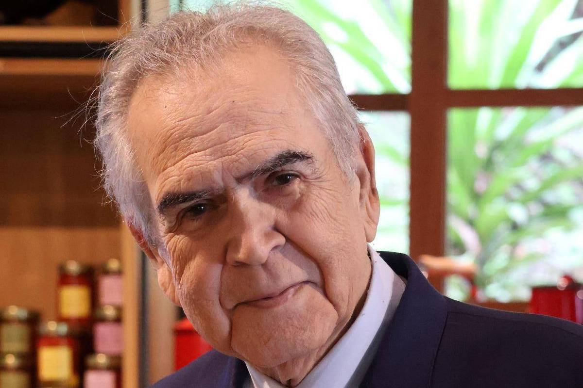 Homem idoso com cabelo grisalho veste terno azul escuro e camisa branca, posando em ambiente interno com prateleiras e janela ao fundo.