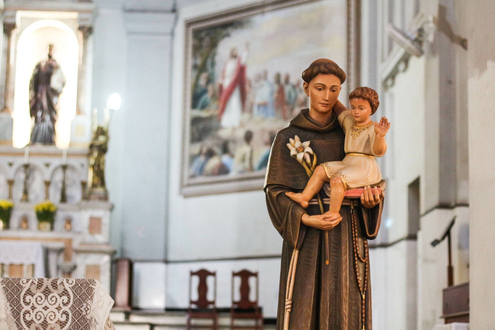 Estátua de santo com hábito marrom segurando criança vestida de branco dentro de igreja. Fundo mostra altar, pintura religiosa e bancos de madeira.