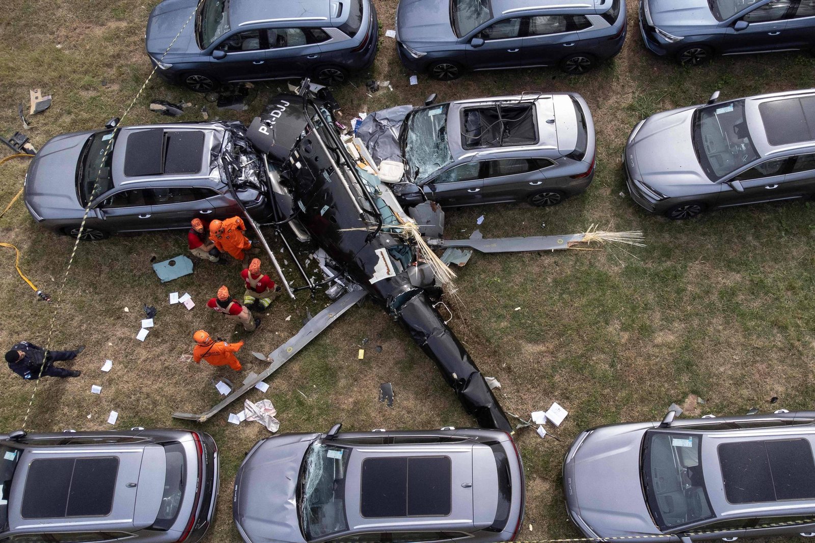 Helicóptero preto da polícia caiu entre carros cinza estacionados em área gramada. Equipe de resgate com uniformes laranja trabalha ao redor da aeronave danificada.