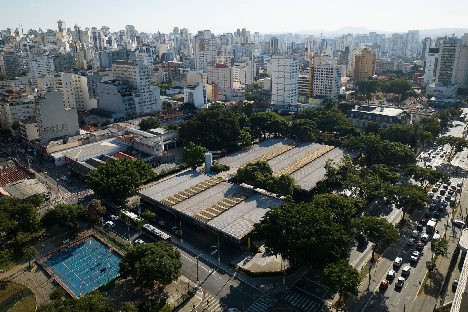 Vista aérea mostra área urbana densa com prédios altos ao fundo e área verde com árvores no centro. Em primeiro plano, quadra esportiva azul e vermelha e ruas com tráfego de veículos.