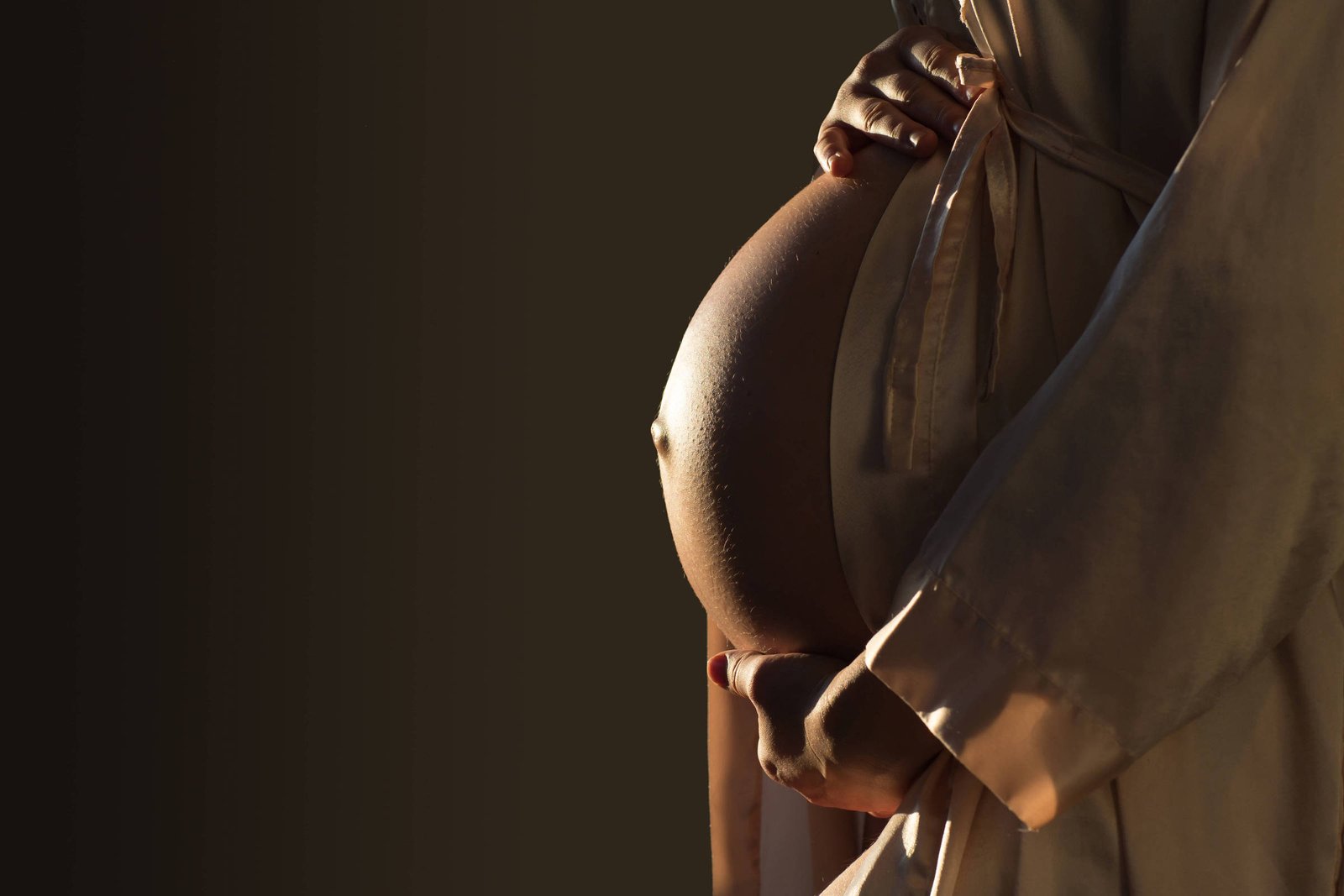 A imagem mostra uma mulher grávida segurando sua barriga com as duas mãos. Ela está vestindo uma roupa clara e a iluminação destaca a forma arredondada da barriga. O fundo é escuro, criando um contraste com a figura da mulher.