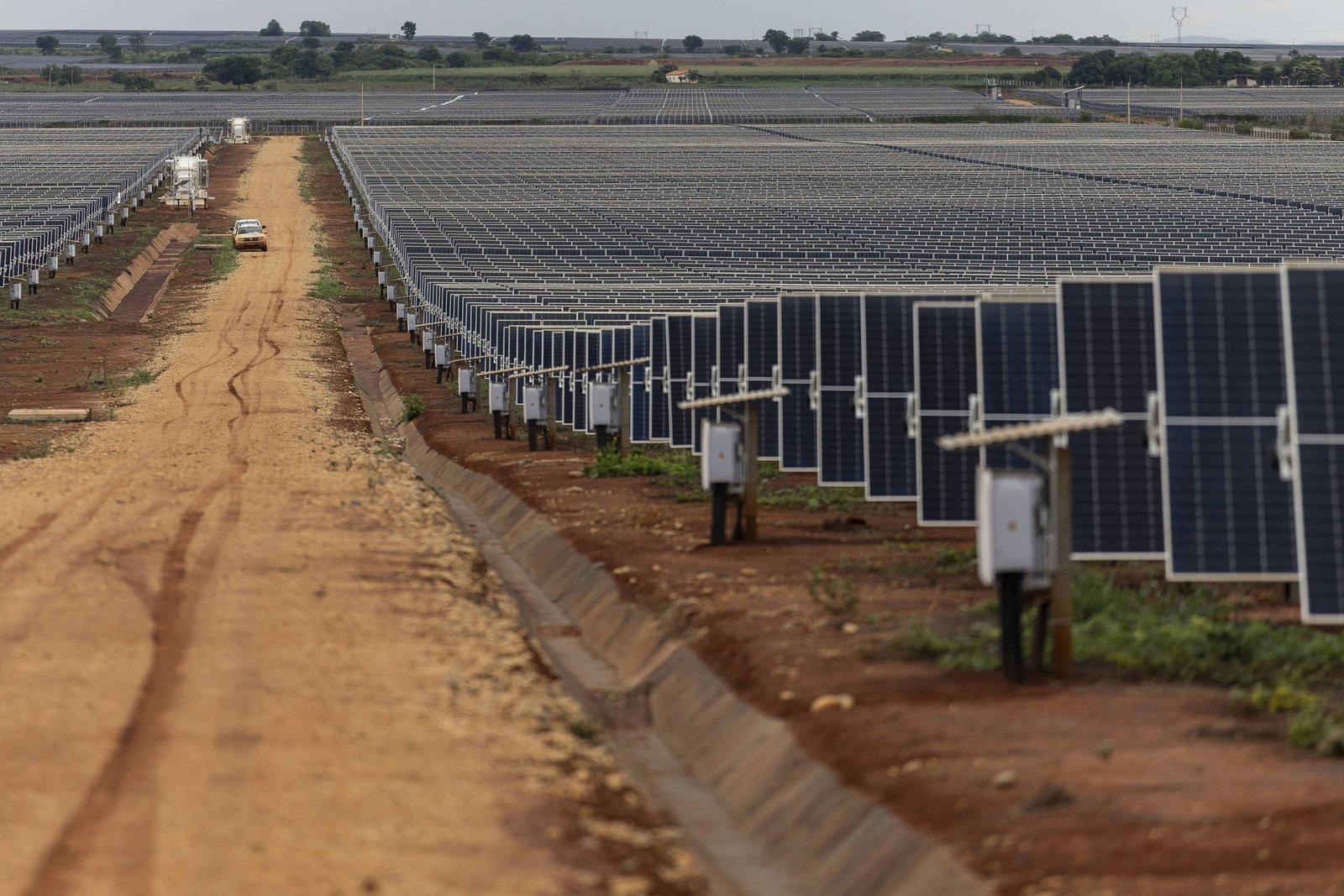 A imagem mostra um grande campo de painéis solares dispostos em fileiras, com um caminho de terra ao centro. O céu está nublado, e ao fundo é possível ver mais painéis solares e algumas construções. A área é predominantemente árida, com vegetação esparsa ao redor dos painéis.