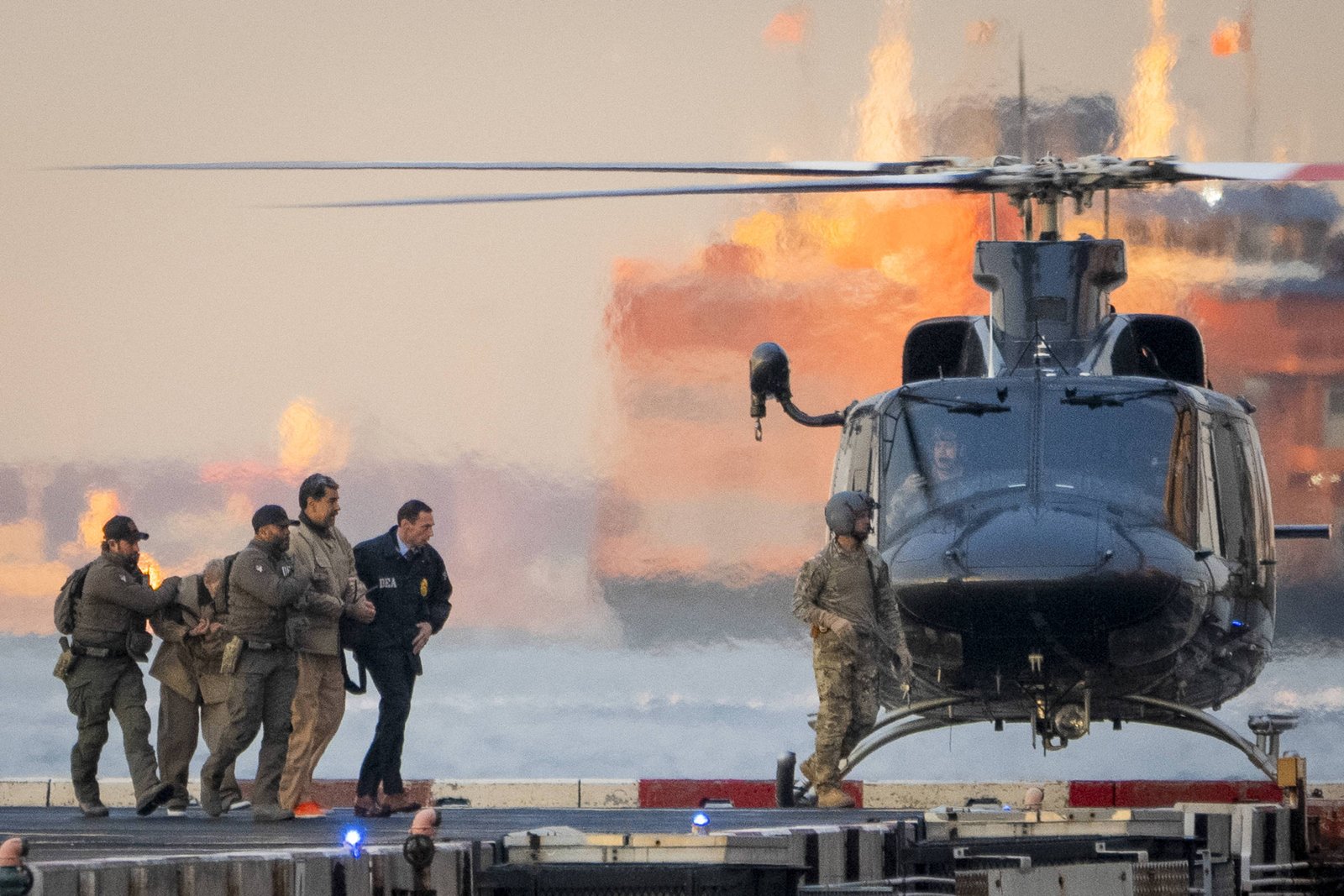 Cinco agentes de segurança escoltam um homem algemado em direção a um helicóptero preto estacionado em um píer próximo ao mar. Ao fundo, há uma grande embarcação com chamas visíveis, indicando atividade industrial ou naval. O cenário é ao entardecer, com luz natural suave e reflexos na água.