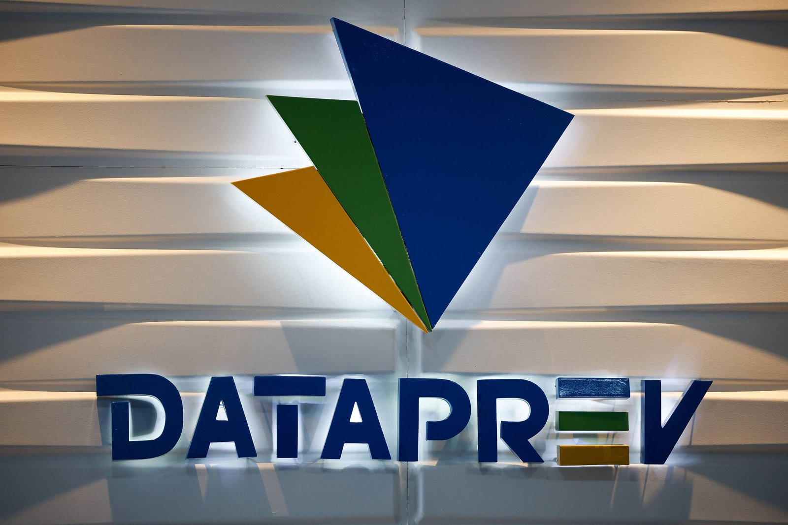 Logotipo da Dataprev com letras azuis em relevo sobre parede branca com iluminação indireta. Acima do nome, três formas triangulares sobrepostas nas cores azul, verde e amarelo.