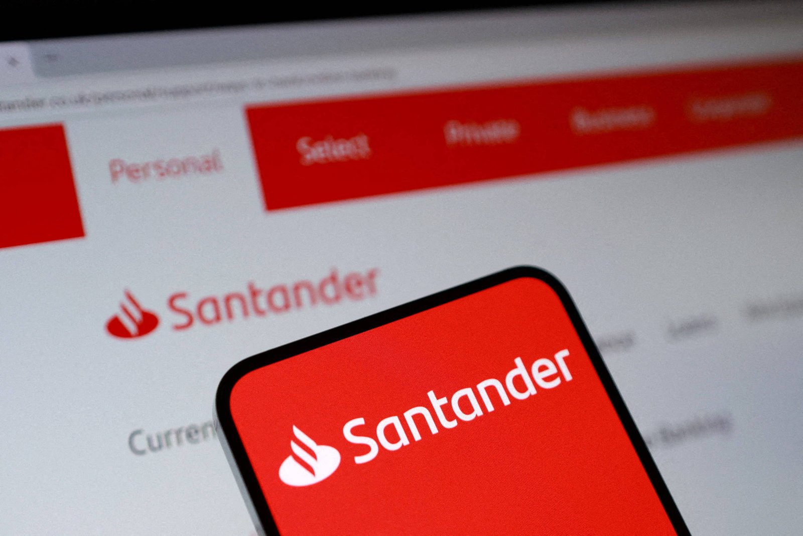 Tela de computador exibe site do Santander com menu em vermelho e texto em branco. Em primeiro plano, celular mostra logotipo branco do Santander sobre fundo vermelho.