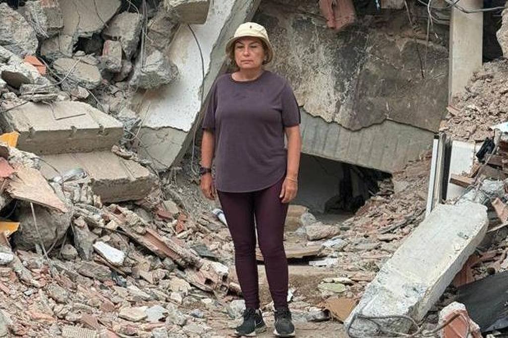 Mulher de camiseta cinza, calça escura e chapéu claro está em meio a escombros de concreto e tijolos de um prédio destruído.