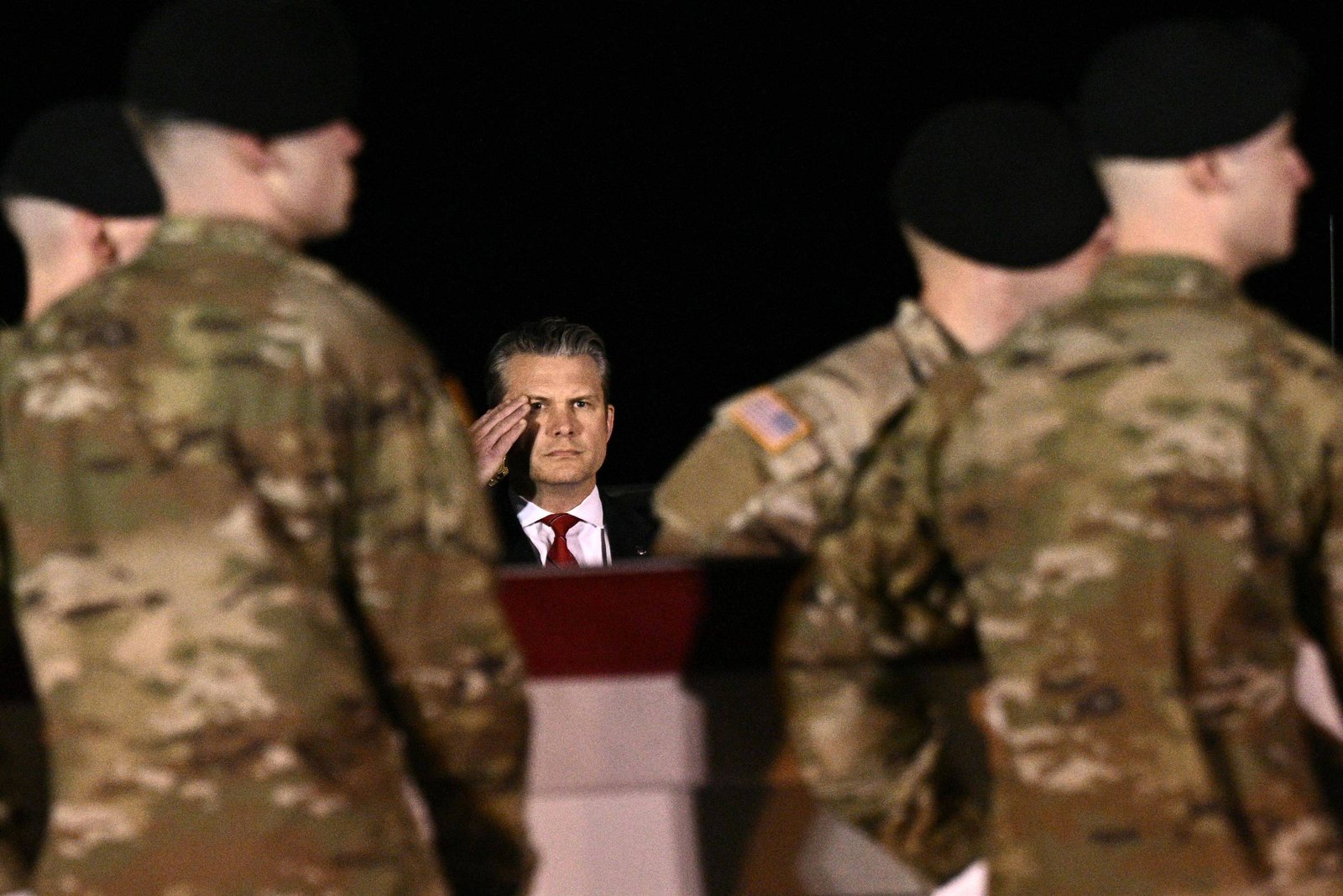 Homem de terno e gravata vermelha faz saudação com a mão direita ao olhar para frente, cercado por quatro soldados com uniformes camuflados e boinas pretas, em ambiente escuro.