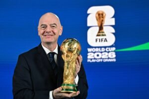 Homem de terno preto segura a taça dourada da Copa do Mundo em frente a fundo azul com logo da FIFA e texto