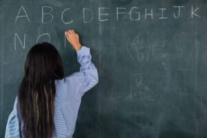 Mulher de costas escreve letras do alfabeto em giz branco no quadro negro de uma sala de aula.