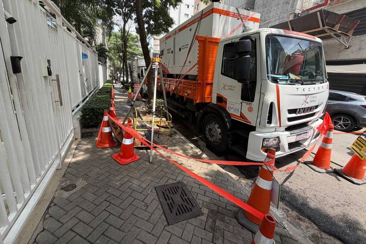 Caminhão branco e laranja estacionado ao lado de calçada bloqueada por cones e fita de segurança vermelha. Equipamento de medição está montado na calçada próxima ao veículo. Edifícios e árvores cercam a área em dia claro.