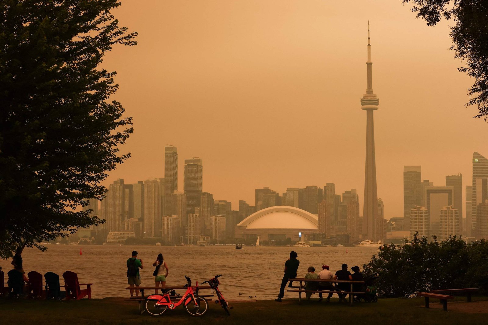 Vista do horizonte de Toronto com a CN Tower e o Rogers Centre ao fundo sob céu alaranjado. Pessoas observam a paisagem em parque à beira do lago, com árvores e bicicletas na frente.