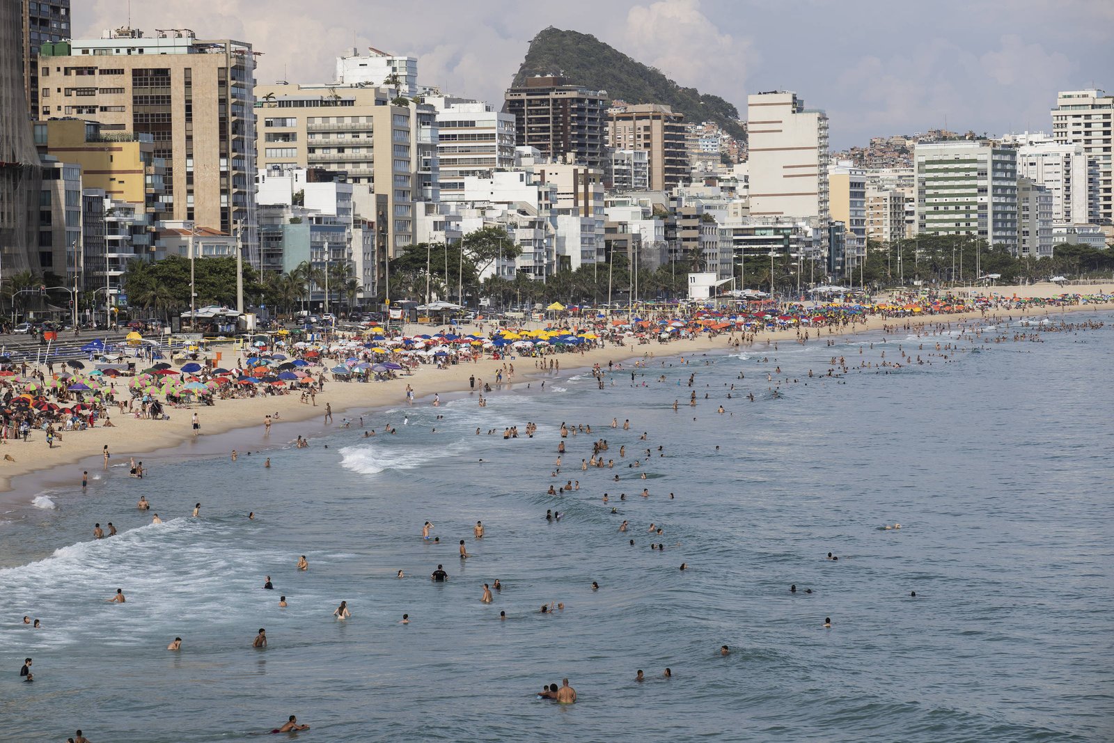 Praia extensa com muitas pessoas na areia sob guarda-sóis coloridos e várias pessoas nadando no mar. Ao fundo, prédios altos e morros cobertos de vegetação sob céu parcialmente nublado.