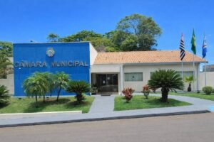 Prédio da Câmara Municipal com parede azul à esquerda e parte branca à direita, rodeado por jardim com palmeiras e arbustos. Três bandeiras estão hasteadas à direita, sob céu azul claro.