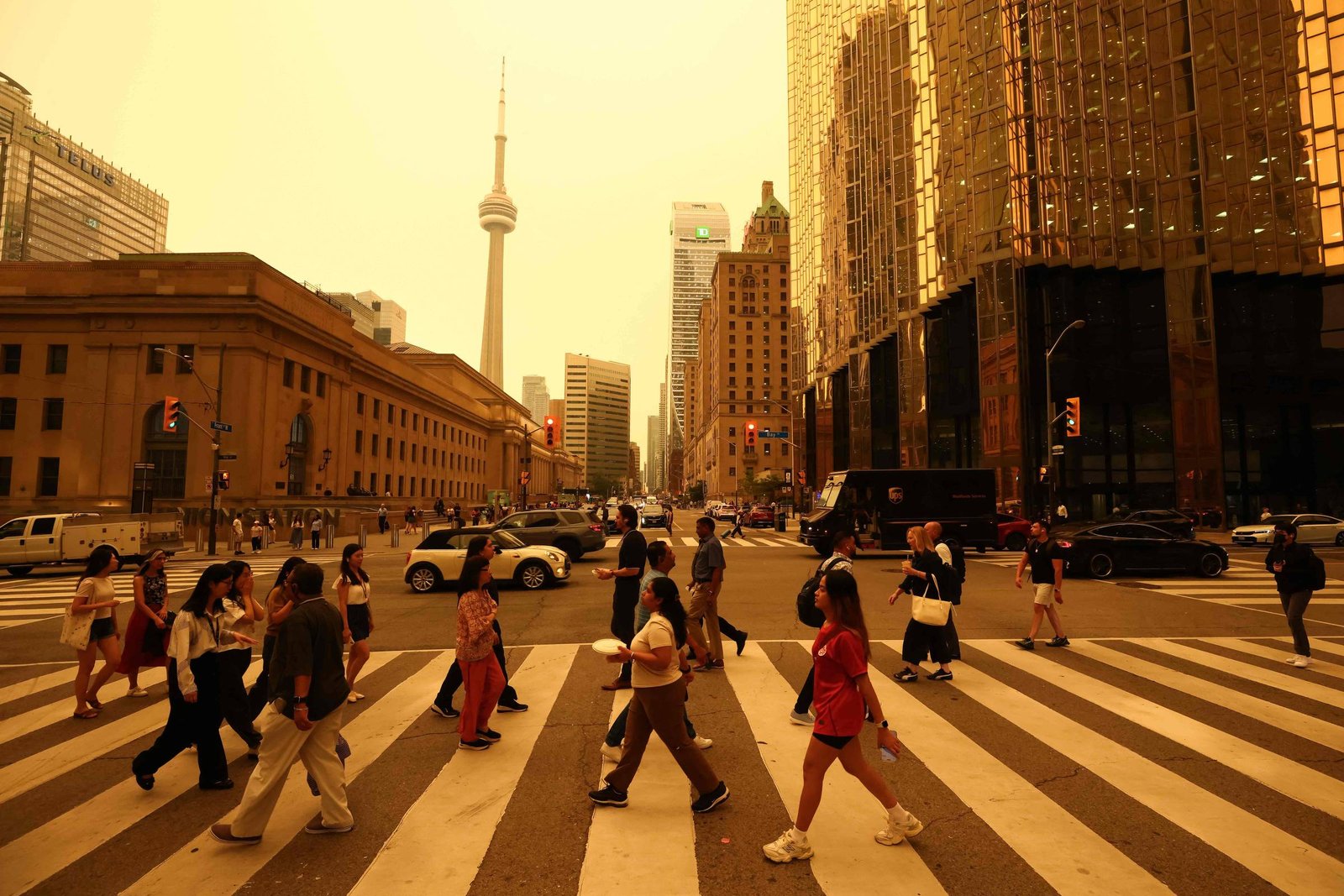 Várias pessoas cruzam a faixa de pedestres em uma avenida movimentada de Toronto, com o CN Tower visível ao fundo. Edifícios altos cercam a rua, e veículos aguardam o sinal de trânsito.