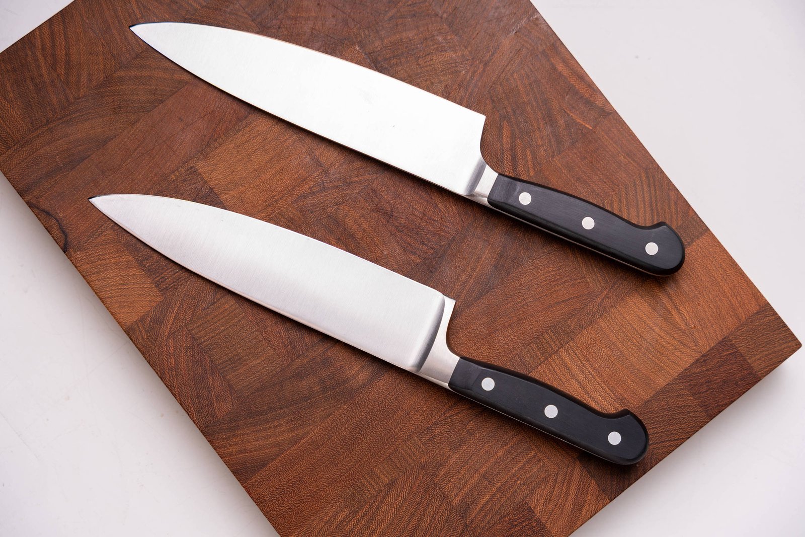 Duas facas de chef com lâminas de aço inoxidável e cabos pretos com três rebites estão posicionadas paralelamente sobre uma tábua de madeira escura.