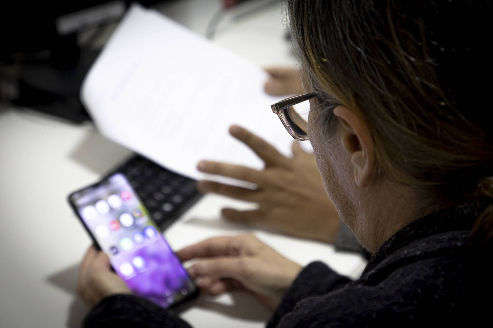 A imagem mostra uma pessoa de costas, usando óculos, focada em um smartphone que está em suas mãos. Ao fundo, há uma mão de outra pessoa apontando para um documento em uma mesa, que também possui um teclado. O ambiente parece ser um escritório.