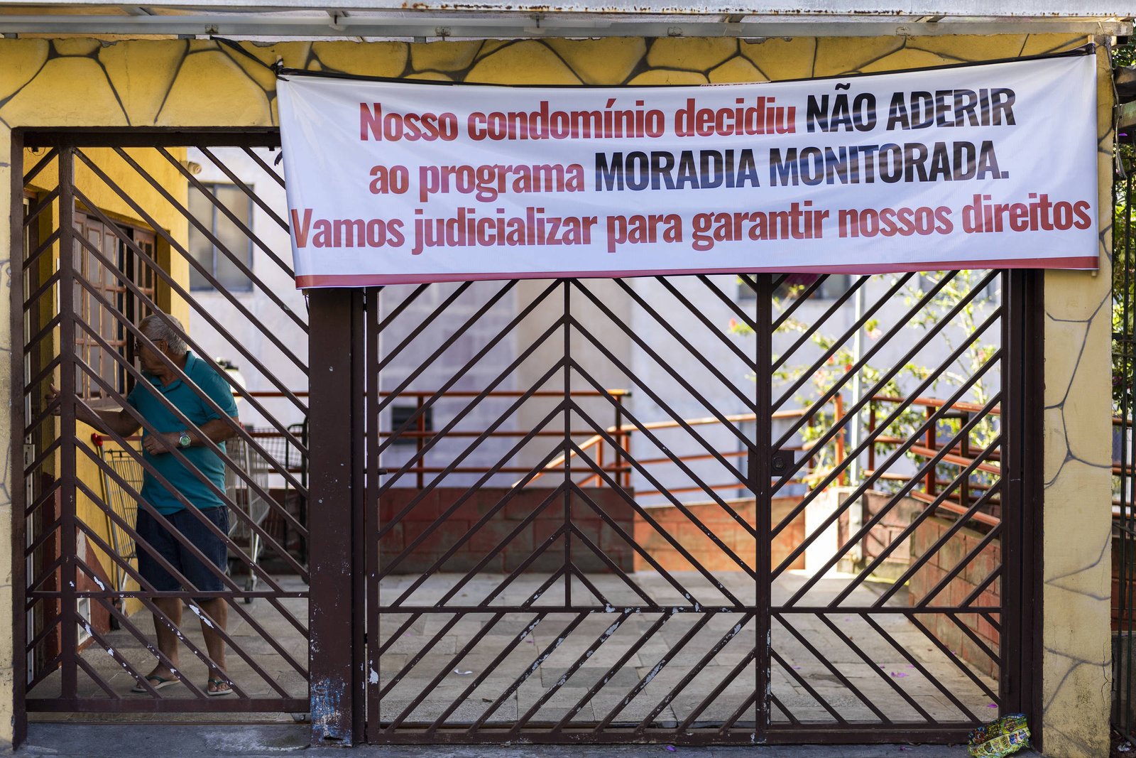 Faixa branca com texto em vermelho e preto pendurada em portão de condomínio residencial. A faixa diz que o condomínio decidiu não aderir ao programa Moradia Monitorada e que judicializará para garantir direitos. Homem está atrás do portão de metal marrom, em frente à entrada do condomínio.