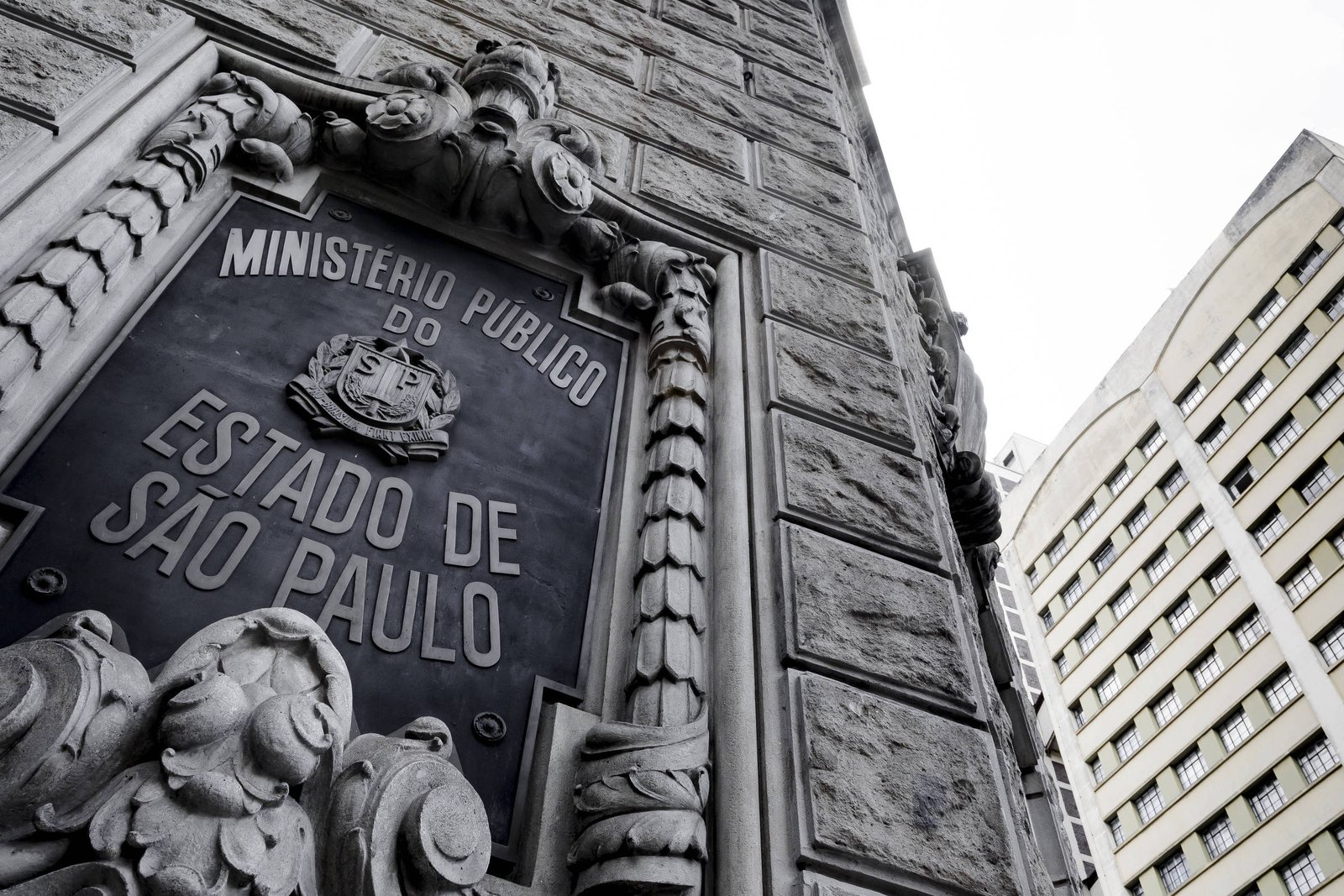 A imagem mostra a fachada do edifício do Ministério Público do Estado de São Paulo. O destaque é uma placa em metal com a inscrição
