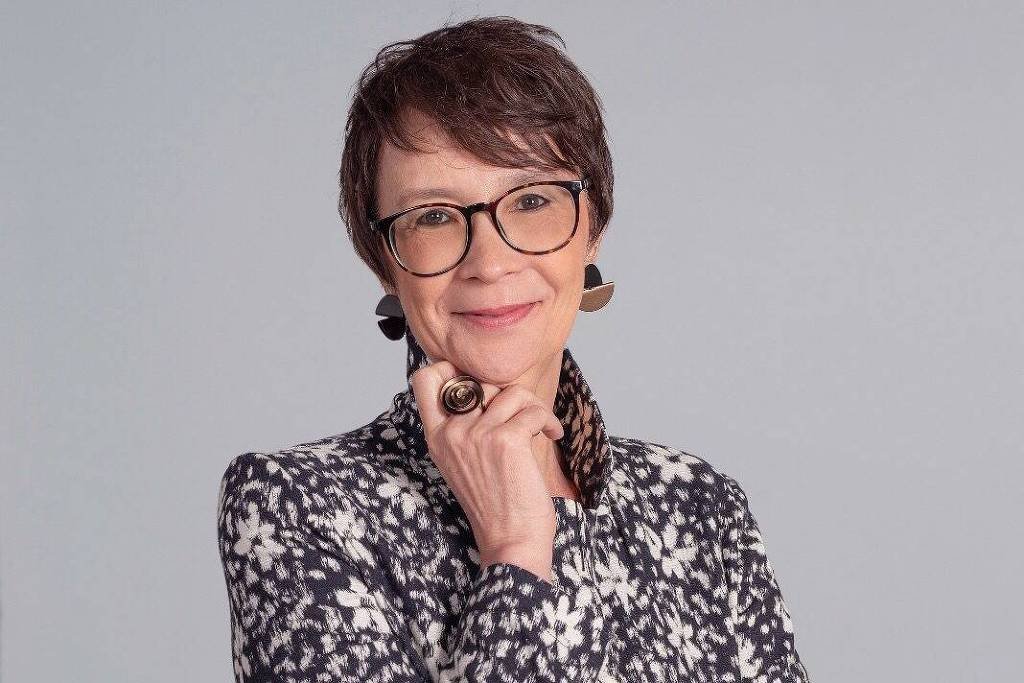 Mulher de cabelos curtos e castanhos usa óculos de armação preta e roupa com estampa preta e branca. Ela apoia a mão direita no queixo, usando anel grande, e sorri levemente. Fundo cinza neutro.