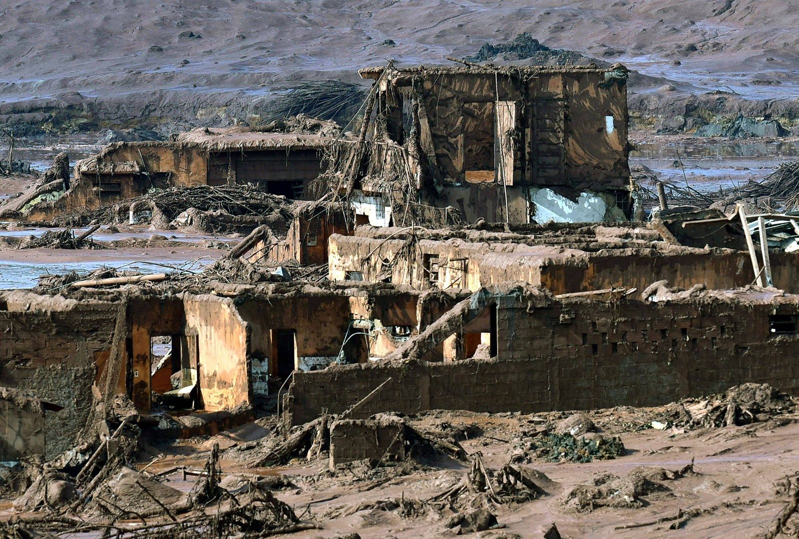 A imagem mostra estruturas em ruínas, com paredes desmoronadas e cobertas por detritos. O cenário é árido, com solo seco e uma aparência de devastação. Não há vegetação visível, e a área parece ter sido severamente afetada por um desastre ambiental.