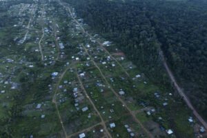 Vista aérea mostra assentamento com casas dispersas em área verde, com estradas de terra delimitando lotes. À direita, floresta densa e estrada de terra margeiam o assentamento.