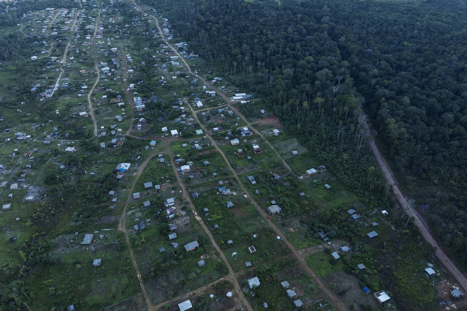 Vista aérea mostra assentamento com casas dispersas em área verde, com estradas de terra delimitando lotes. À direita, floresta densa e estrada de terra margeiam o assentamento.