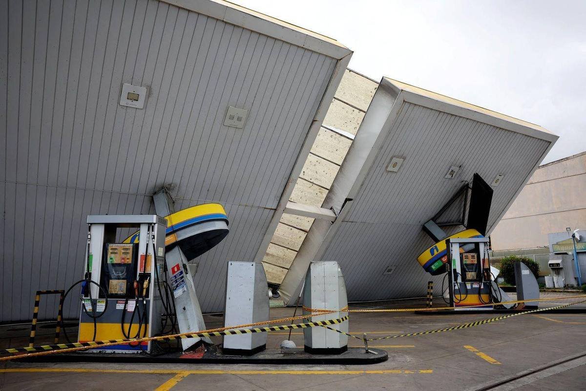Cobertura metálica de posto de gasolina desabada sobre as bombas de combustível. Área isolada com fita amarela de segurança, sem pessoas presentes.