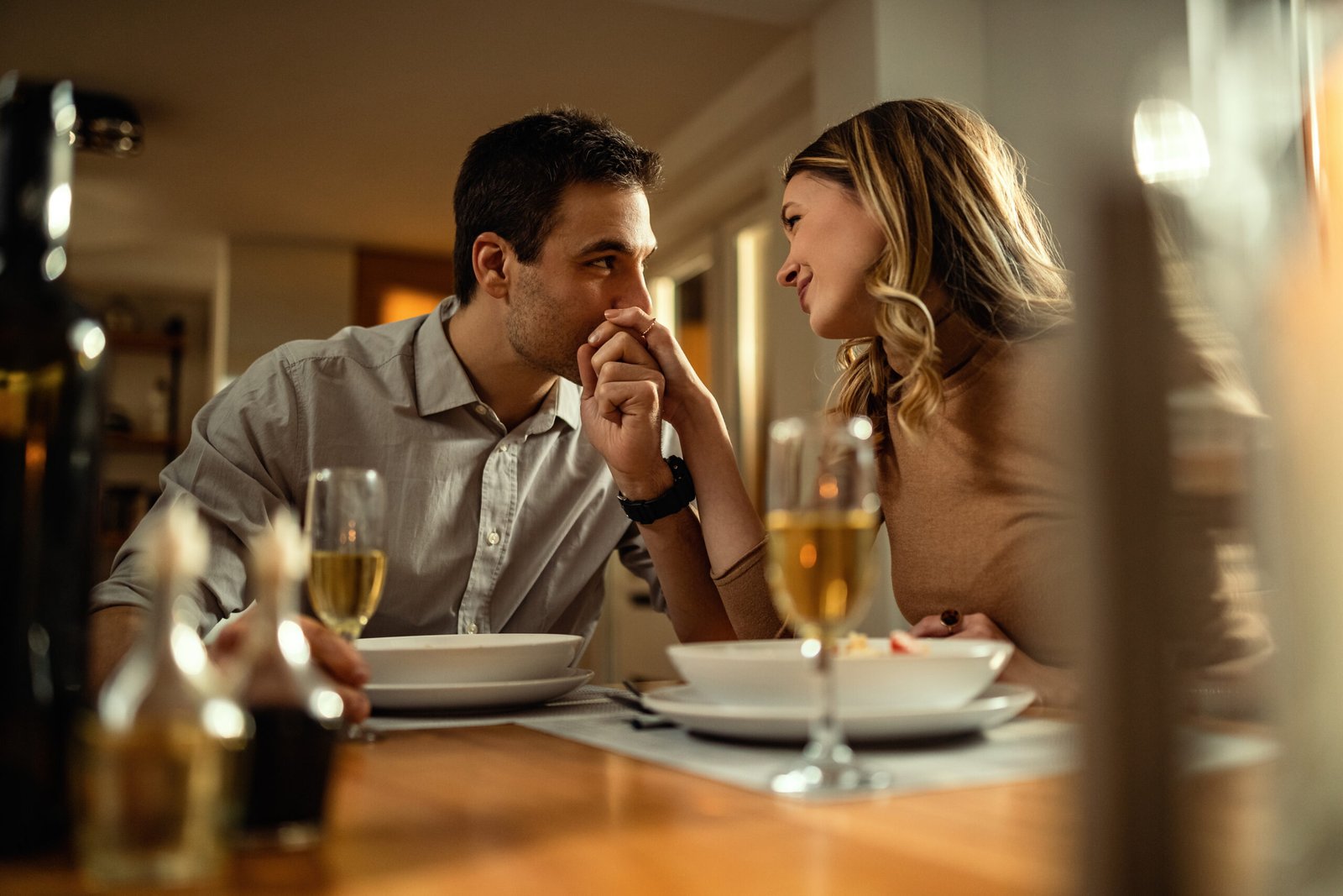 Happy woman enjoying at dining table while being kissed in a hand by her boyfriend.