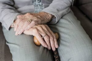 A imagem mostra as mãos de uma pessoa idosa descansando sobre as pernas. As mãos são enrugadas e uma delas segura um bastão de madeira. A pessoa está vestindo uma blusa cinza e as calças são de tom claro. Um anel de ouro e um anel com pedra são visíveis em uma das mãos.