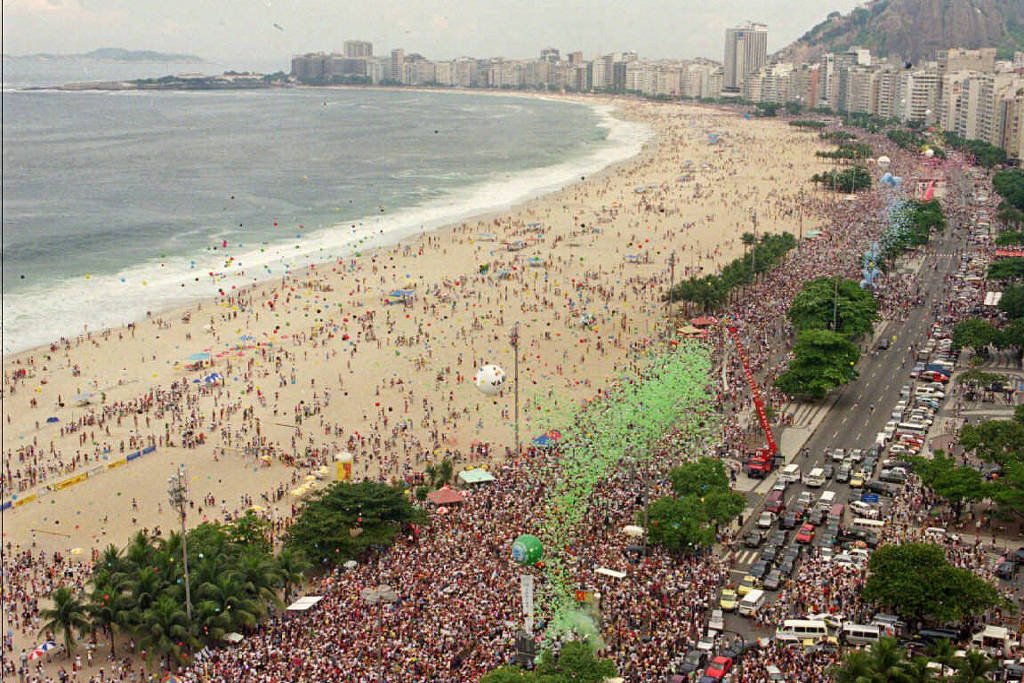 Grande concentração de pessoas ocupa a avenida e a faixa de areia da praia de Copacabana, no Rio de Janeiro, durante evento diurno. Edifícios altos e o Morro do Leme aparecem ao fundo, com o mar à esquerda e trânsito intenso na via paralela à praia.