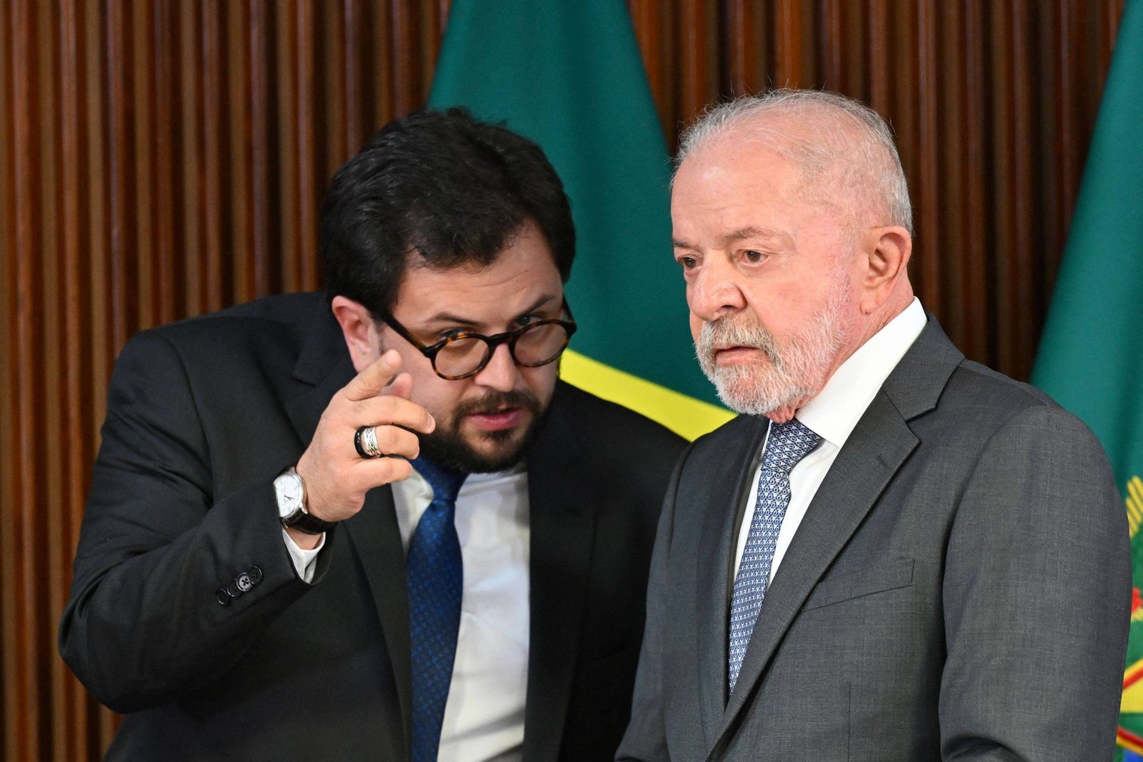 Dois homens de terno conversam em ambiente formal com bandeira do Brasil ao fundo. O homem à esquerda, Marco Aurélio, é branco, tem cabelos castanhos curtos e lisos e barba castanha, usa óculos e faz gesto com a mão direita; o homem à direita, Lula, é branco, tem cabelos e barba grisalhos e curtos e observa atentamente.