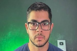 Homem jovem de cabelo curto e barba aparada usa óculos de armação preta e camiseta azul escura. Ele está posicionado centralmente contra um fundo cinza com iluminação verde à esquerda e um troféu do YouTube desfocado à direita.